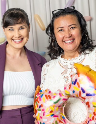Two women standing in front of a sprinkle backdrop holding a donut balloon